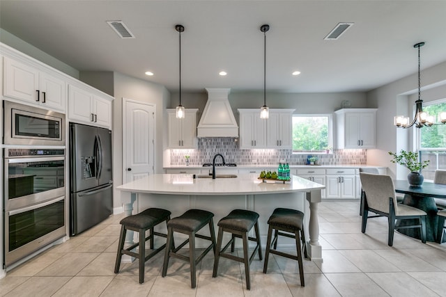 kitchen with custom exhaust hood, light countertops, appliances with stainless steel finishes, a kitchen island with sink, and white cabinetry