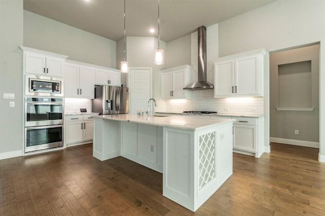 kitchen featuring appliances with stainless steel finishes, pendant lighting, white cabinets, a kitchen island with sink, and wall chimney exhaust hood