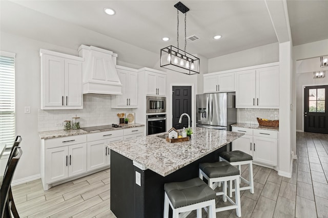 kitchen featuring premium range hood, white cabinets, hanging light fixtures, an island with sink, and appliances with stainless steel finishes