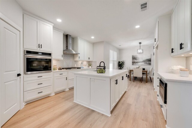 kitchen with appliances with stainless steel finishes, a kitchen island with sink, wall chimney range hood, light hardwood / wood-style floors, and white cabinetry