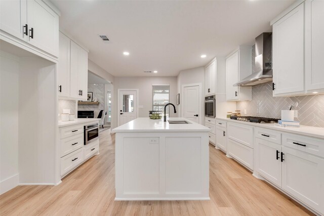 kitchen featuring light wood-type flooring, stainless steel appliances, wall chimney range hood, white cabinets, and an island with sink