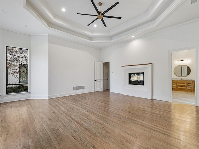 unfurnished living room featuring ceiling fan, a multi sided fireplace, a raised ceiling, light hardwood / wood-style flooring, and ornamental molding