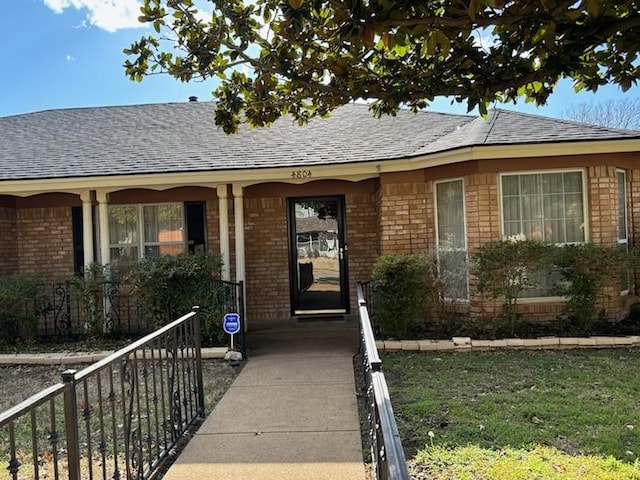 doorway to property featuring fence, brick siding, and roof with shingles