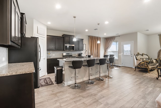 kitchen with a center island with sink, hanging light fixtures, light wood-type flooring, appliances with stainless steel finishes, and light stone counters