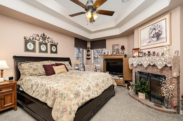 carpeted bedroom featuring ceiling fan, a tray ceiling, and a brick fireplace