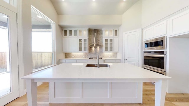 kitchen with sink, wall chimney exhaust hood, an island with sink, white cabinets, and appliances with stainless steel finishes