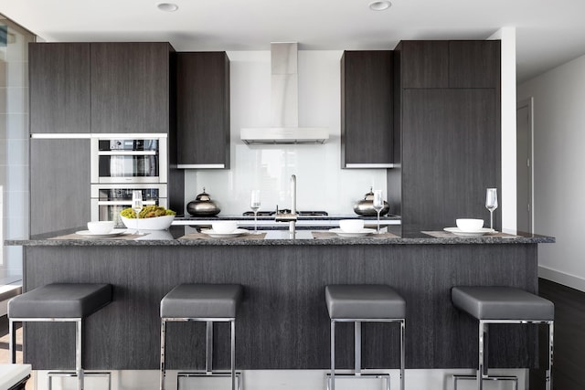 kitchen with a breakfast bar area, hardwood / wood-style flooring, wall chimney range hood, and appliances with stainless steel finishes