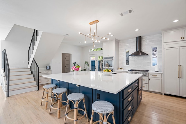 kitchen with wall chimney range hood, hanging light fixtures, stainless steel appliances, a spacious island, and white cabinets