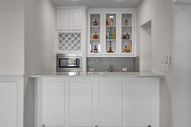 bar with backsplash, stainless steel microwave, white cabinets, and light stone counters