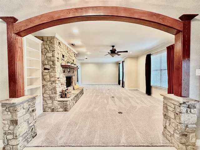 unfurnished living room with light carpet, a stone fireplace, ceiling fan, and crown molding