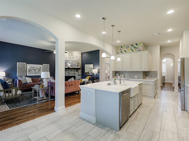 kitchen with ceiling fan, stainless steel appliances, an island with sink, decorative light fixtures, and a fireplace