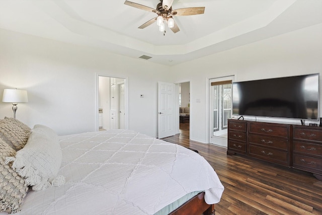 bedroom with ceiling fan, ensuite bathroom, visible vents, a tray ceiling, and dark wood finished floors