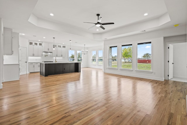 unfurnished living room with a tray ceiling, plenty of natural light, ceiling fan with notable chandelier, and light wood-type flooring