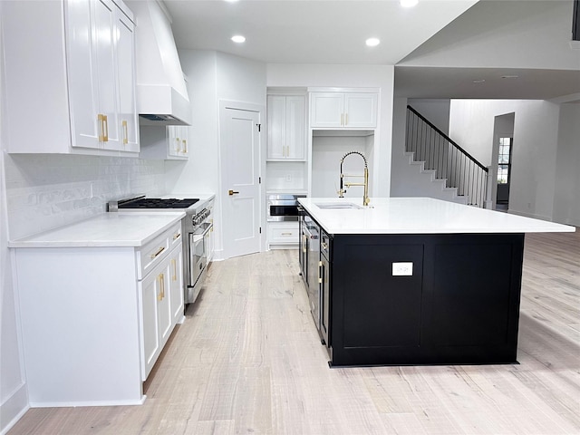 kitchen with appliances with stainless steel finishes, premium range hood, white cabinetry, and an island with sink