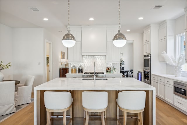 kitchen featuring custom exhaust hood, white cabinets, light wood-type flooring, an island with sink, and appliances with stainless steel finishes