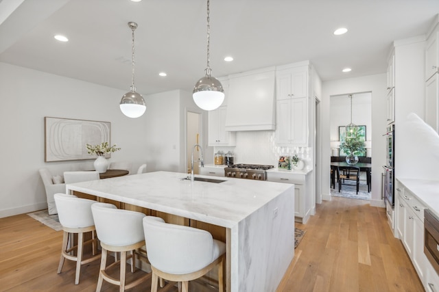 kitchen with premium range hood, sink, decorative light fixtures, a center island with sink, and white cabinets