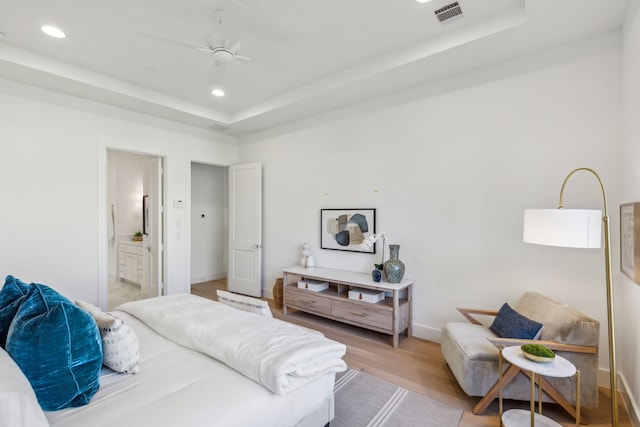 bedroom with ceiling fan, light hardwood / wood-style floors, a raised ceiling, and ensuite bath