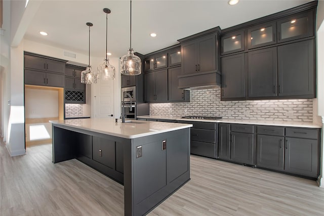 kitchen with decorative light fixtures, a center island with sink, appliances with stainless steel finishes, custom range hood, and light hardwood / wood-style floors