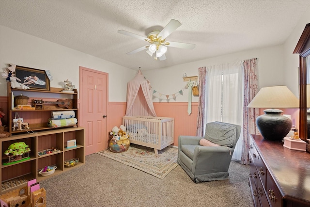 carpeted bedroom featuring a nursery area, a wainscoted wall, ceiling fan, and a textured ceiling