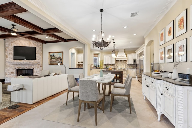 tiled dining room featuring coffered ceiling, a stone fireplace, beamed ceiling, ceiling fan with notable chandelier, and ornamental molding