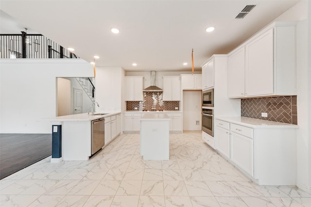 kitchen featuring white cabinets, an island with sink, wall chimney exhaust hood, appliances with stainless steel finishes, and light countertops