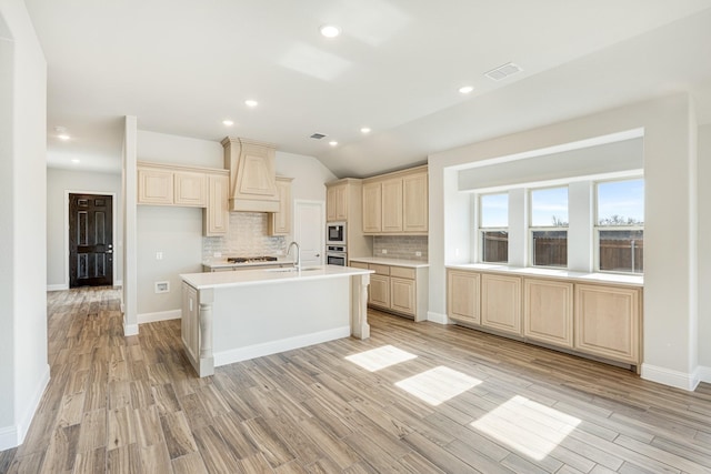 kitchen featuring custom exhaust hood, an island with sink, light wood-type flooring, backsplash, and sink