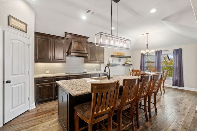 kitchen with sink, a kitchen island with sink, dark brown cabinets, custom range hood, and a kitchen bar