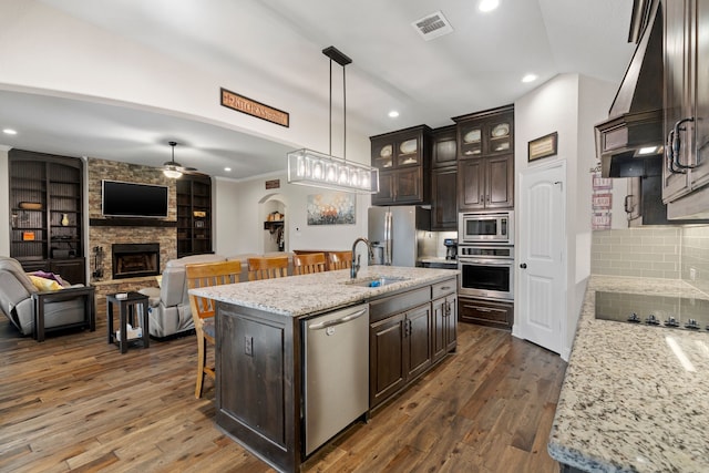 kitchen with appliances with stainless steel finishes, a stone fireplace, a kitchen island with sink, and dark brown cabinets