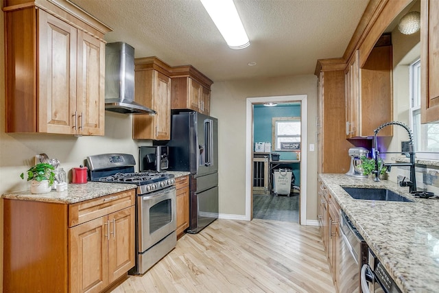 kitchen with light wood-type flooring, sink, light stone counters, appliances with stainless steel finishes, and wall chimney range hood