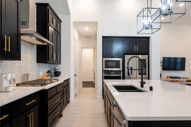 kitchen with wall chimney exhaust hood, stainless steel appliances, light hardwood / wood-style floors, decorative light fixtures, and a kitchen island with sink