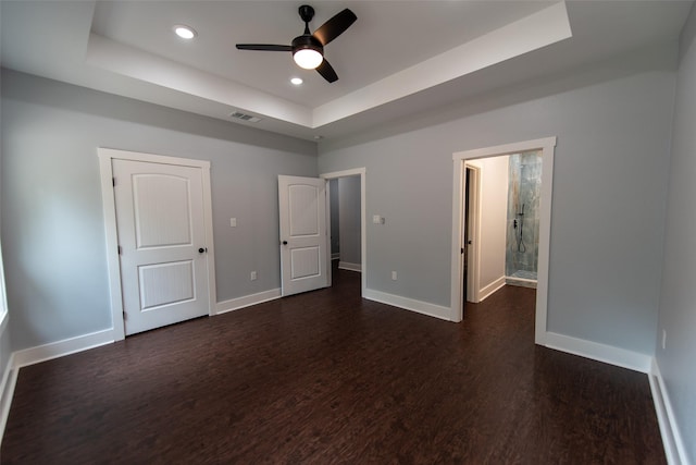 unfurnished bedroom with a tray ceiling, ensuite bath, ceiling fan, and dark wood-type flooring