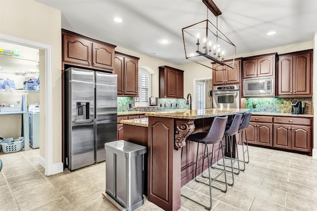 kitchen featuring backsplash, a kitchen island with sink, light stone countertops, a notable chandelier, and stainless steel appliances