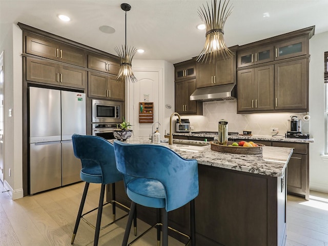 kitchen with extractor fan, appliances with stainless steel finishes, light stone counters, dark brown cabinets, and a center island with sink