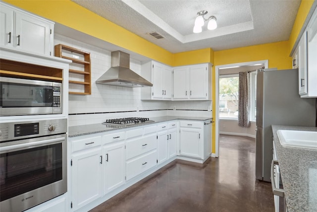 kitchen with stainless steel appliances, a raised ceiling, wall chimney range hood, and white cabinets