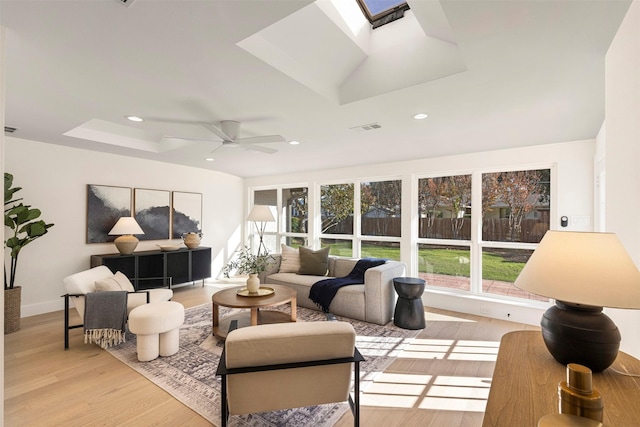 living room featuring a skylight, light hardwood / wood-style floors, a raised ceiling, and ceiling fan