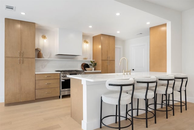 kitchen featuring premium range hood, sink, stainless steel range, light hardwood / wood-style floors, and a kitchen bar