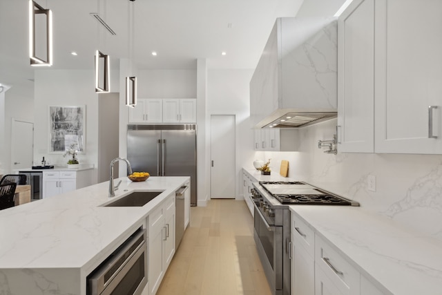 kitchen with pendant lighting, high end appliances, white cabinets, wall chimney range hood, and sink