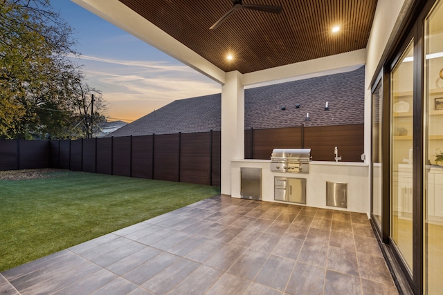 patio terrace at dusk with area for grilling, ceiling fan, a yard, and exterior kitchen