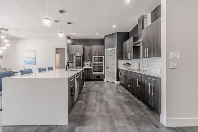 kitchen featuring stainless steel appliances, hanging light fixtures, wall chimney exhaust hood, and an island with sink