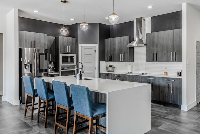 kitchen with wall chimney exhaust hood, stainless steel appliances, an island with sink, decorative light fixtures, and a breakfast bar