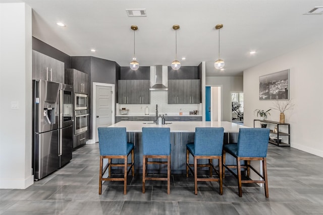 kitchen featuring a center island with sink, wall chimney range hood, hanging light fixtures, and appliances with stainless steel finishes