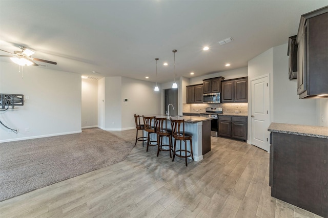 kitchen with dark brown cabinetry, light hardwood / wood-style floors, decorative backsplash, a center island with sink, and appliances with stainless steel finishes