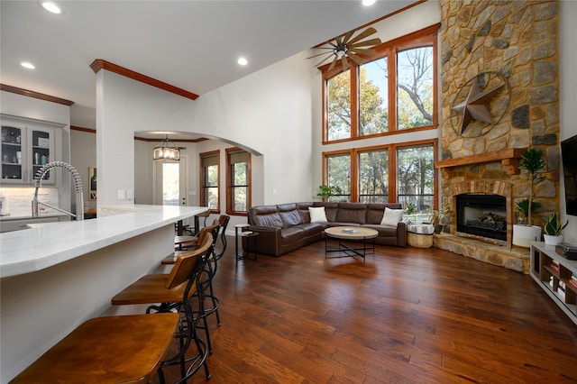 living area featuring arched walkways, a stone fireplace, ceiling fan with notable chandelier, dark wood finished floors, and crown molding