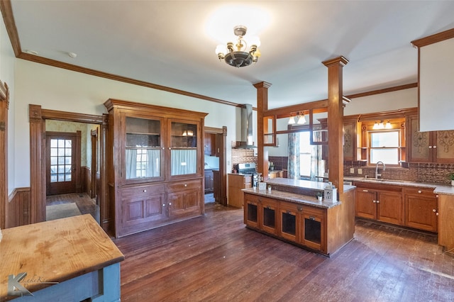 kitchen with dark wood-type flooring, wall chimney exhaust hood, sink, crown molding, and decorative columns