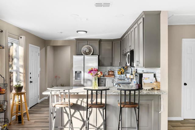kitchen with appliances with stainless steel finishes, a kitchen breakfast bar, light stone countertops, hardwood / wood-style floors, and backsplash