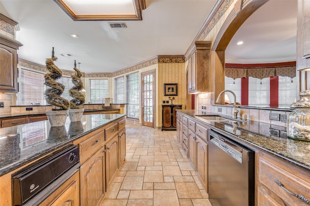 kitchen featuring sink, backsplash, dark stone counters, stainless steel dishwasher, and crown molding