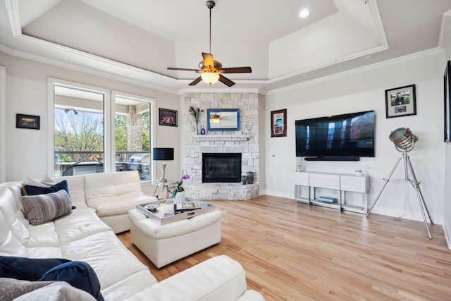 living room with a raised ceiling, ceiling fan, crown molding, light hardwood / wood-style flooring, and a stone fireplace