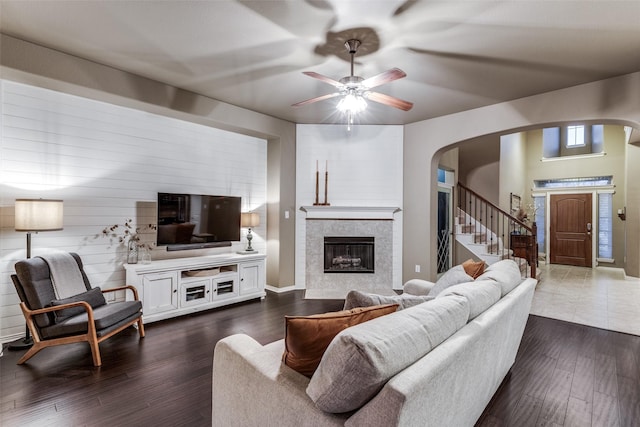 living area with ceiling fan, dark wood finished floors, stairs, a tile fireplace, and arched walkways