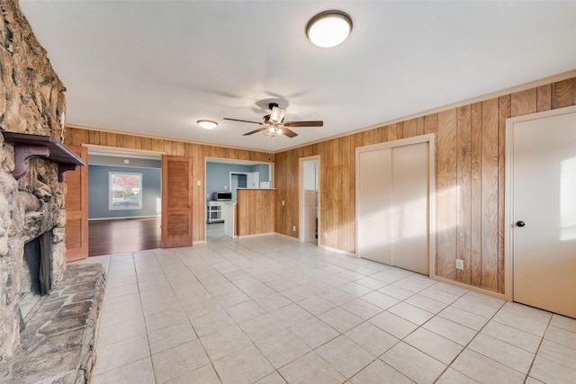 unfurnished living room with ceiling fan, wood walls, a stone fireplace, and light tile patterned floors