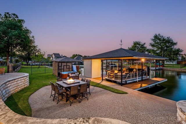 patio terrace at dusk with a boat dock, a water view, an outdoor fire pit, and a lawn